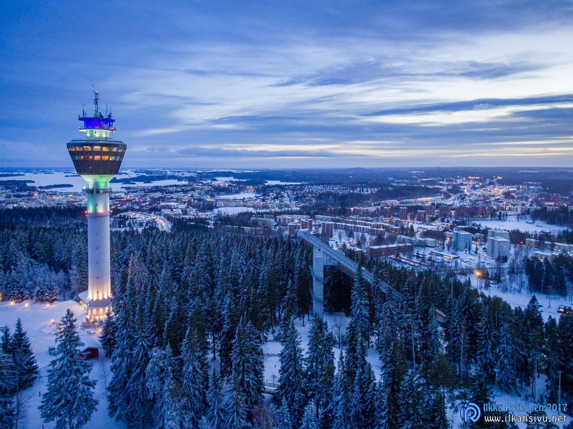 An illuminated tower rises above a snowy forest in blue twilight