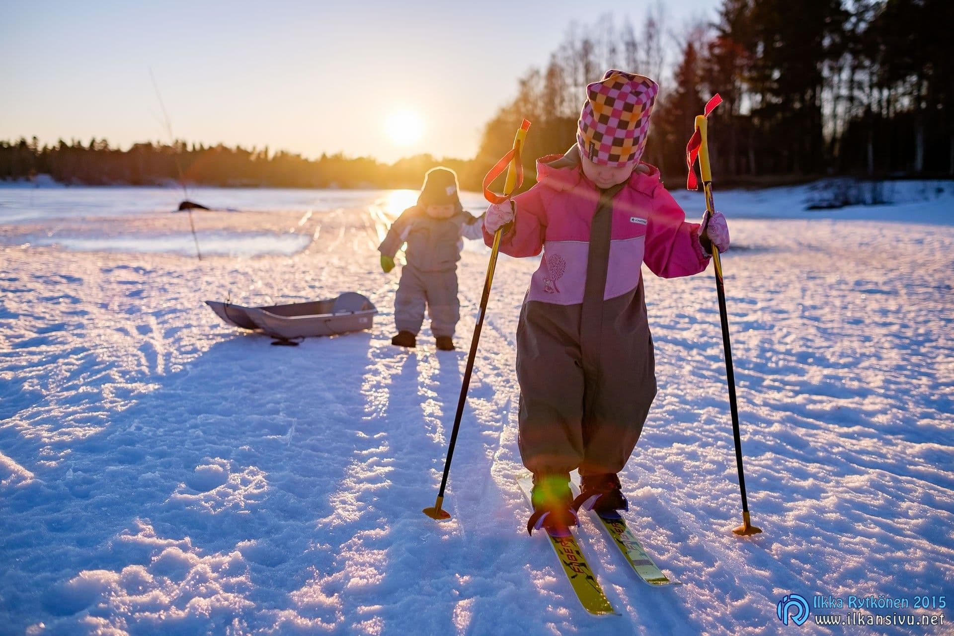 Lapset lumessa matalan auringon vastavalossa, säädetyn kuvan etuala ja vaatteet näkyvät vaaleampina