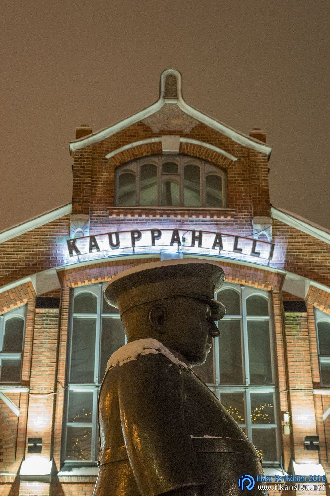 Patsas valaistun kauppahallin edessä yökuvassa, jossa varjojen yksityiskohdat erottuvat vaaleampina