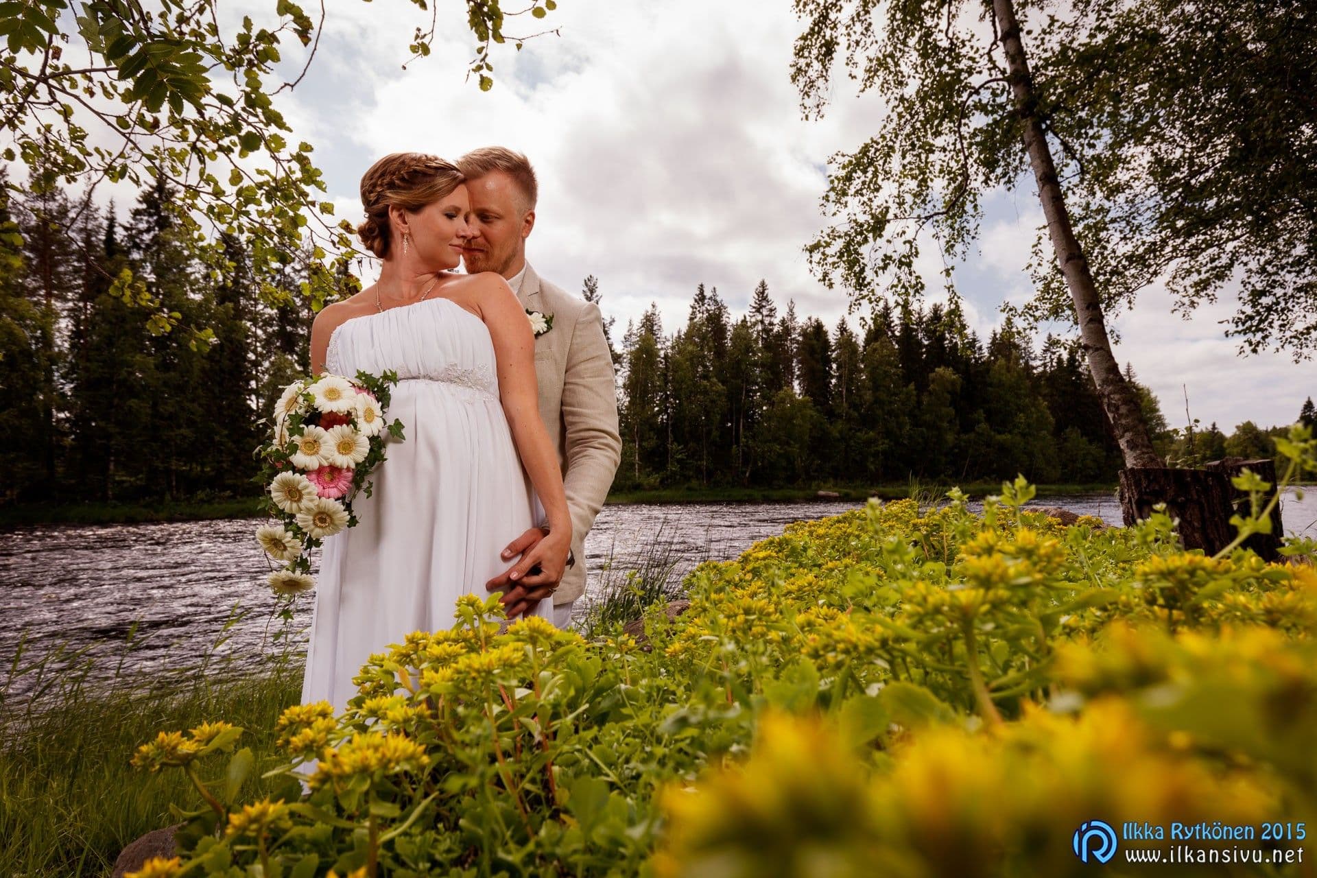 Laura and Jukka stand close together on the bank of the Kiiminkijoki river among yellow flowers