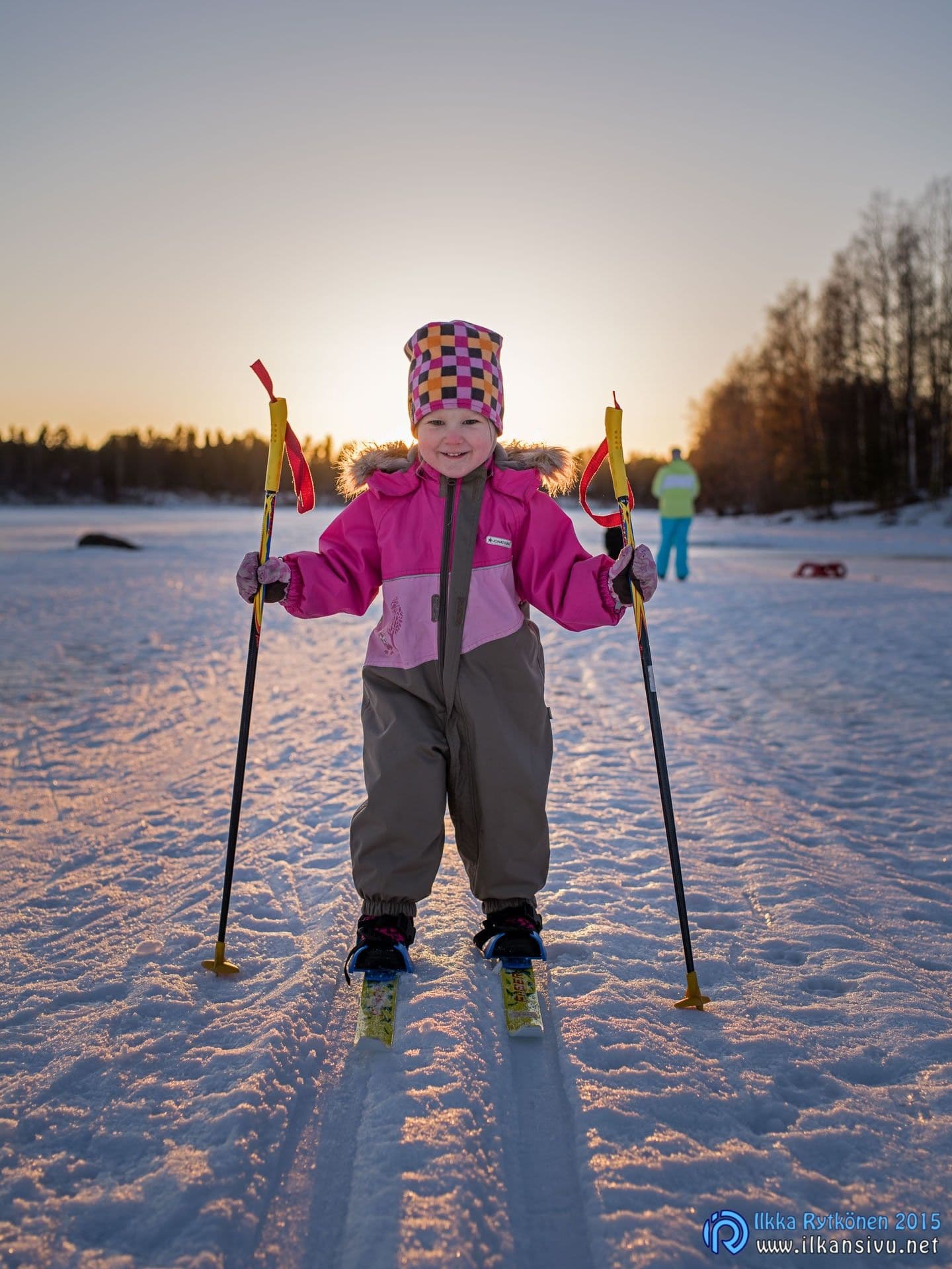 Lapsi seisoo suksilla lumessa vastavalossa, säädetyssä kuvassa kasvot ja vaatetus erottuvat vaaleampina